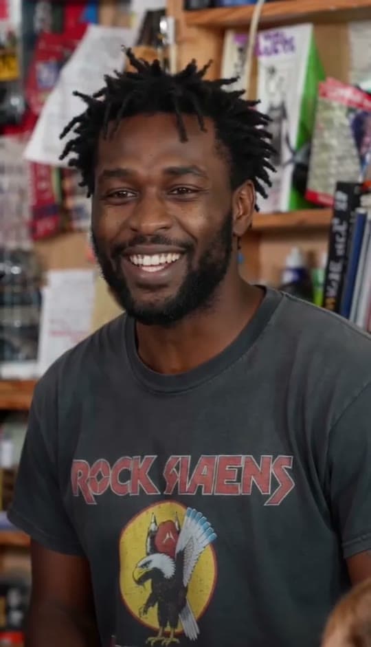 The lead singer seat in a Tiny Desk AI sample video: a smiling man with short dreadlocks in a grey graphic tee